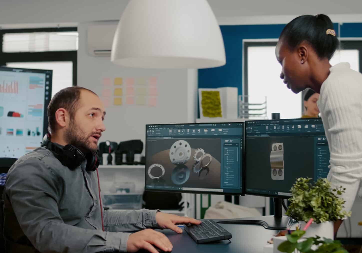 Mechanical engineer working on computer designing in CAD software 3D model of engine while african coworker asking help. Man using pc with two monitors innovating motor prototype in creative agency